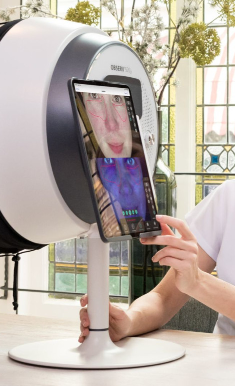 Person som håller en surfplatta som visar ansiktsdata, framför en hudvårdsanalysenhet i ett ljust rum med målat glasfönster.