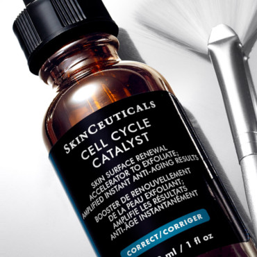 En flaska märkt "SkinCeuticals Cell Cycle Catalyst" på en reflekterande yta, bredvid ett metalliskt hudvårdsverktyg.